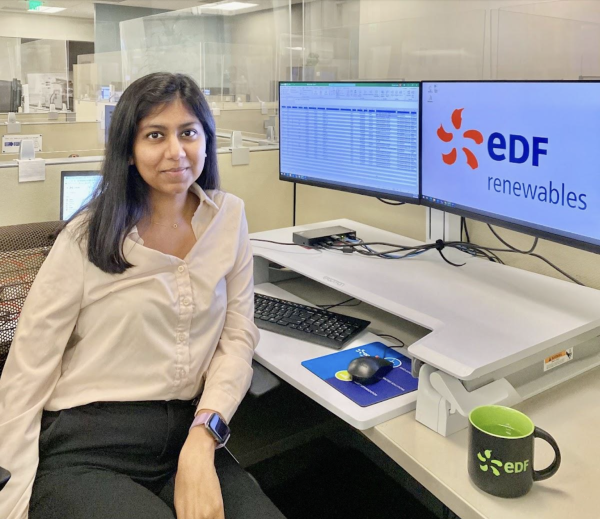 A woman sits in a cubicle in an office in front of two computer monitors, one with a spreadsheet and one with the company logo visible. She is smiling