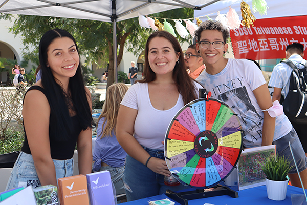 Three students from the SDSU Active Minds club smile at a table. Brochures on self-confidence and overcoming procrastination as well as a multi-colored chance wheel are on the table in front of them