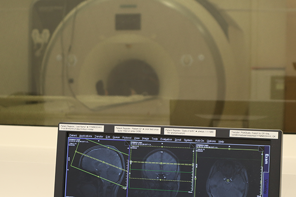Brain scans on a computer in the foreground; in the background, through a window, a person is lying inside the opening of an MRI machine