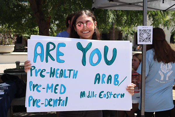 A smiling student wearing sunglasses holding a sign that says "Are you pre-health, pre-med, pre-dental, Arab/Middle-Eastern" in teal letters