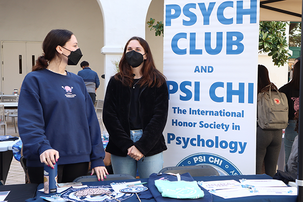 Two students stand at a table with fun brain-related probs with a banner that says Psych Club and Psi Chi