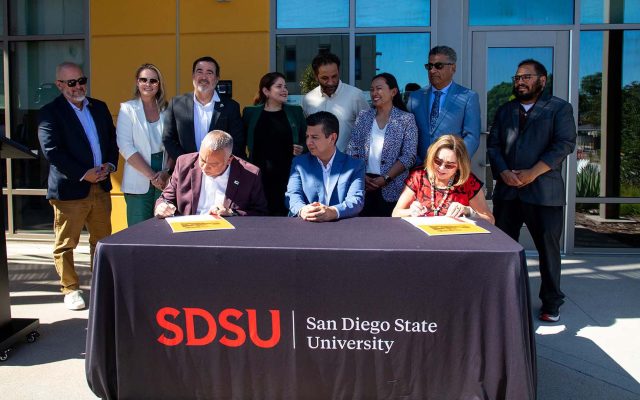 Mark Sanches, David Alvarez, and Adela del ta Torre outside, sitting at desk with table cloth with SDSU | San Diego State University on it, with 8 other people standing behind.